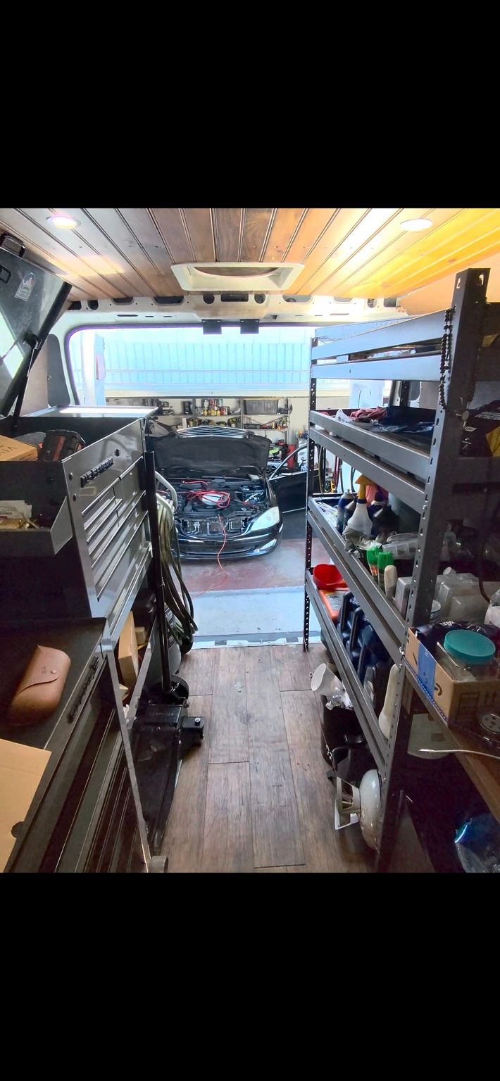 Inside a mobile service van with a tool chest and parts shelving, looking out at a black sedan with its hood open